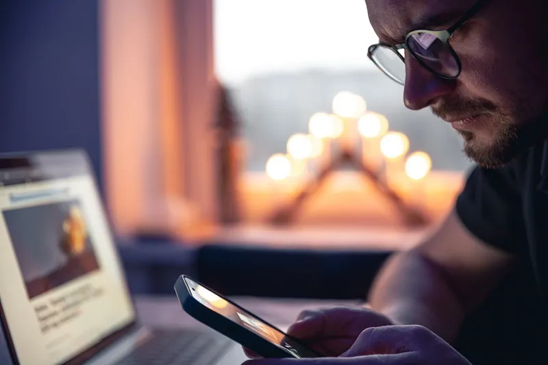 Ein Mann mit einem Smartphone sitzt spät in der Nacht vor einem Laptop.