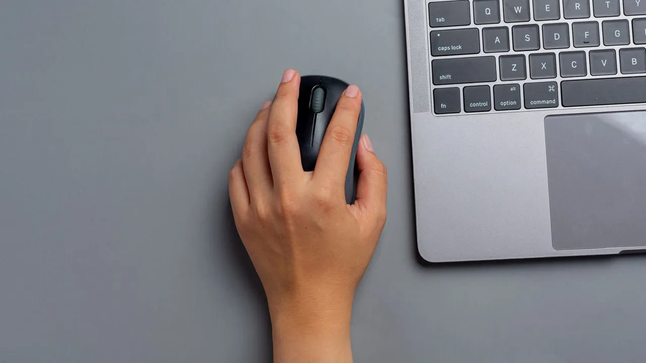 A hand holding a wireless mouse next to a laptop