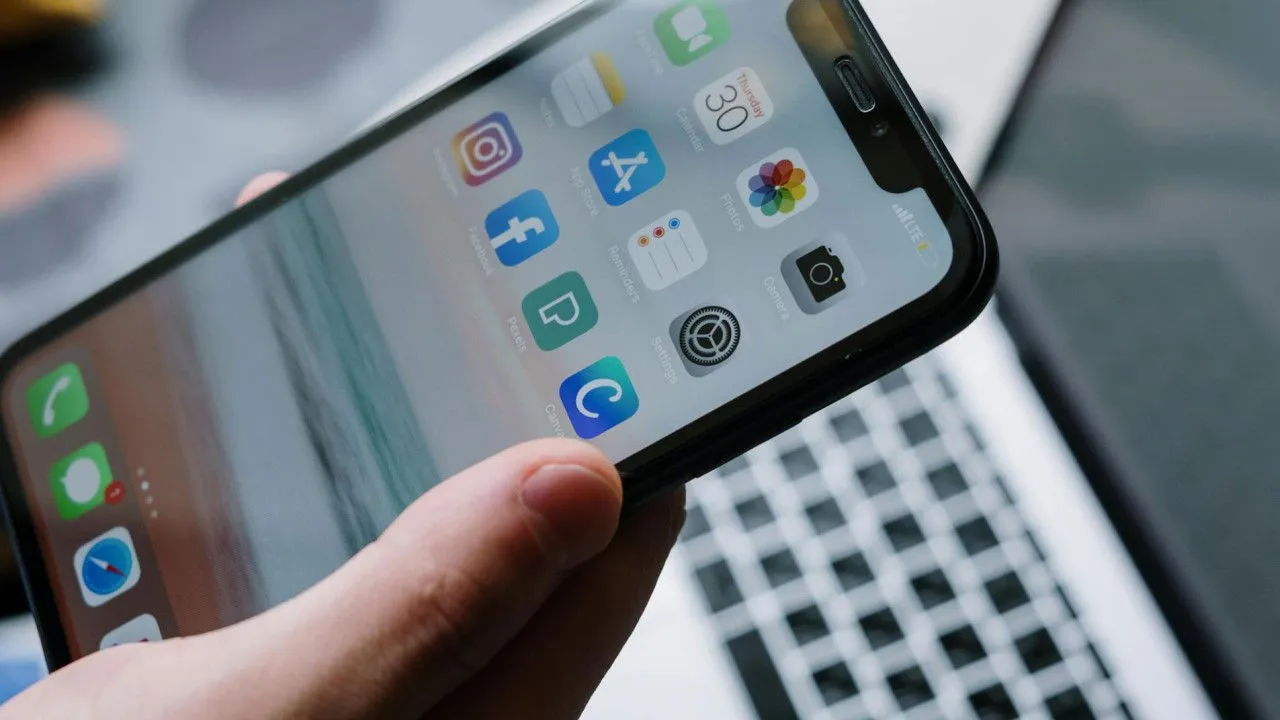 A hand holding a smartphone in front of an open laptop.