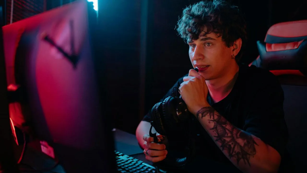 A Man in a Black Shirt Talking on a Headphone's Mic While Looking at a Computer Monitor.